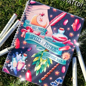 May include: A laminated spiral-bound coloring book titled "Spells & Potions" with a fantasy theme. The cover features illustrations of crystals, potions, a wand, and a crescent moon. White markers are scattered around the book on a grassy surface.