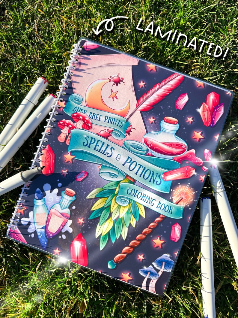 May include: A laminated spiral-bound coloring book titled "Spells & Potions" with a fantasy theme. The cover features illustrations of crystals, potions, a wand, and a crescent moon. White markers are scattered around the book on a grassy surface.
