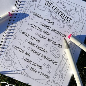 May include: A spiral-bound notebook with a checklist titled "THE CHECKLIST" in black ink. The checklist includes items like "Bubbling Potions" and "Crystal Ball." Two white markers with pink and blue tips are on the page. The notebook is on a grassy surface.