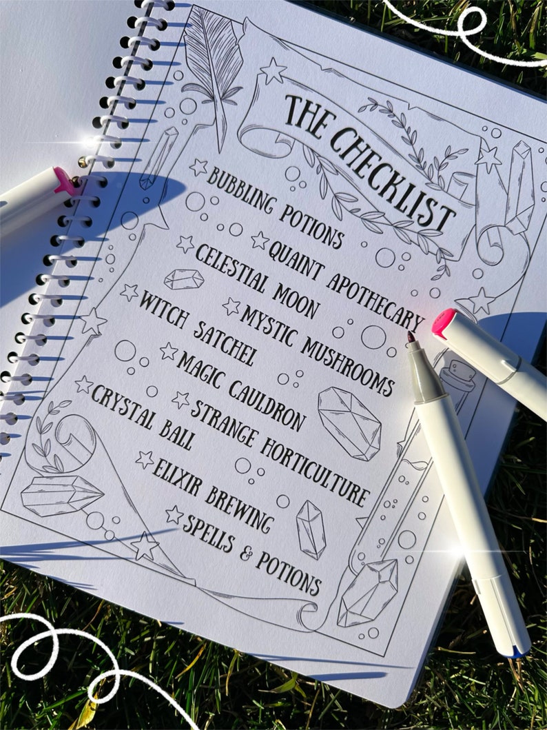 May include: A spiral-bound notebook with a checklist titled "THE CHECKLIST" in black ink. The checklist includes items like "Bubbling Potions" and "Crystal Ball." Two white markers with pink and blue tips are on the page. The notebook is on a grassy surface.