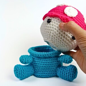 May include: A crocheted toy shaped like a mushroom with a removable top. The mushroom is white with a pink cap and black eyes. The bottom of the mushroom is blue.