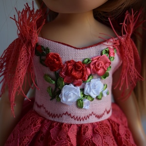 May include: A doll in a pink dress with red and white floral embroidery. The dress has a lace skirt and fringed shoulder accents. The bodice is decorated with red and white roses and green leaves.