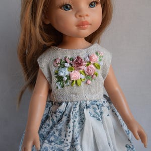 Könnte beinhalten: Puppe mit blauen Augen und blonden Haaren, die ein ärmelloses Kleid trägt. Das Oberteil des Kleides ist hellgrau mit einem gestickten Blumendesign in Rosa, Blau und Burgunder. Der Rock ist weiß mit einem blauen Blumenmuster.