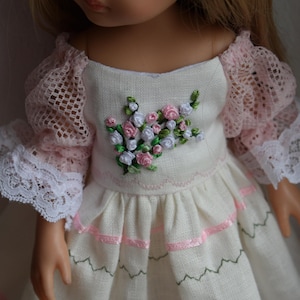 Könnte beinhalten: Eine Puppe in einem cremefarbenen Kleid mit rosa Spitzenärmeln und weißer Spitzenborte. Das Kleid hat eine Blumenstickerei auf dem Oberteil und rosa Bandakzente. Der Rock hat ein Rüschendesign mit Zickzackstich.