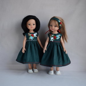 Könnte beinhalten: Zwei Puppen tragen passende grüne Kleider mit gestickten Blumendetails. Die Kleider haben einen gewellten Saum und sind mit roten Nähten verziert. Eine Puppe trägt ein Stirnband mit einer roten Schleife.