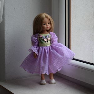 Könnte beinhalten: Puppe mit langen blonden Haaren, die ein lavendelfarbenes Kleid mit floralem Oberteil und ausgestelltem Rock trägt. Das Kleid hat lange Ärmel und wird mit weißen Schuhen kombiniert. Die Puppe steht auf einer Fensterbank.