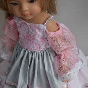 Könnte beinhalten: Eine Puppe in einem rosa-grauen Kleid mit Spitzärmeln. Das Oberteil ist weiß mit einem rosa Blumenmuster. Die Ärmel sind aus rosa Spitze und der Rock ist grau. Eine Perlenkette schmückt die Schulter der Puppe.