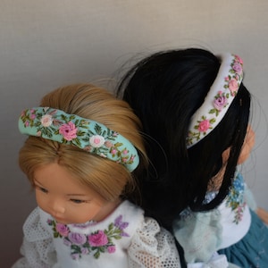 Könnte beinhalten: Zwei Puppen tragen floral bestickte Stirnbänder. Das Stirnband der linken Puppe ist hellblau mit rosa und weißen Blumen. Das Stirnband der rechten Puppe ist weiß mit rosa Blumen.