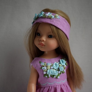 Könnte beinhalten: Eine Puppe mit langem blondem Haar, blauen Augen und einem lavendelfarbenen Kleid. Das Kleid ist mit hellblauer und grüner Blumenstickerei verziert. Ein passendes lavendelfarbenes Stirnband mit floralen Akzenten vervollständigt den Look. Neutraler Hintergrund.
