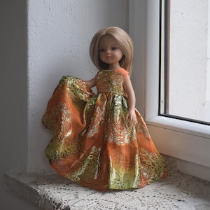 Könnte beinhalten: Eine Puppe mit blonden Haaren in einem orange-grünen gemusterten Kleid mit goldenen Akzenten. Das Kleid hat einen fließenden Rock und ein ärmelloses Oberteil. Die Puppe befindet sich in der Nähe eines Fensters.