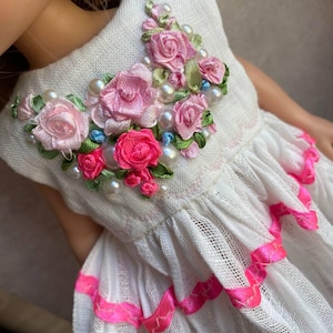 Op de afbeelding: Een witte jurk met een lijfje versierd met roze lintrozen, groene geborduurde bladeren en kleine witte kunstparels. De rok van de jurk is gemaakt van een doorschijnende witte stof met horizontale rijen van felroze lintafwerking.