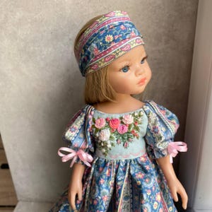 May include: A doll in a blue floral dress and matching headband. The dress has embroidered flowers on the bodice and pink ribbon bows. The doll has blonde hair and blue eyes.