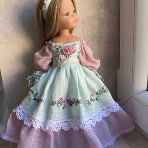 Puede incluir: Muñeca rubia de ojos azules, vestida con un vestido verde claro y rosa. El vestido presenta rosas rosas bordadas y encaje blanco. Se lleva una diadema floral a juego. La muñeca está cerca de una ventana.