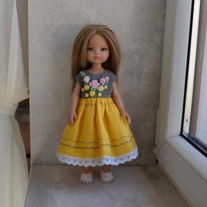 Könnte beinhalten: Eine Puppe in einem gelben Kleid mit einem grauen Oberteil, das mit rosa und gelben Blumen bestickt ist. Das Kleid hat einen weißen Spitzensaum und wird mit weißen Schuhen getragen. Die Puppe hat lange, hellbraune Haare.