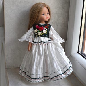 Könnte beinhalten: Eine Puppe mit langem hellbraunem Haar trägt ein weißes Kleid mit Spitzenärmeln und einem dunkelgrünen Oberteil, das mit gestickten roten, rosa und weißen Blumen verziert ist. Der Rock aus weißem, strukturiertem Stoff hat Zickzack-Borten.