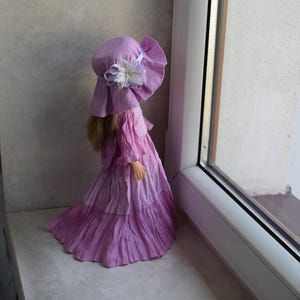 Könnte beinhalten: Eine Puppe in einem lavendelfarbenen Kleid und Hut mit einer weißen Blumenverzierung. Die Puppe steht an einem Fenster, wobei der mehrlagige Rock und der breitkrempige Hut hervorgehoben werden.