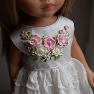 May include: A doll in a white dress with a floral design. The dress has a bodice decorated with pink and white roses and green leaves. The skirt is layered and ruffled. The doll has blonde hair. The image is a close-up.