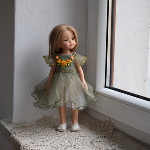May include: A doll with blonde hair wearing a green dress with a floral neckline and a layered skirt. The doll is wearing white shoes and is standing on a lace doily. The dress has yellow accents.