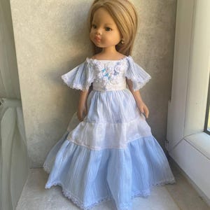 Könnte beinhalten: Eine Puppe mit langem blonden Haar trägt ein gestuftes Kleid mit weißem Oberteil, blauer Blumenstickerei, kurzen Rüschenärmeln und einem Rock aus abwechselnd weißem und hellblauem Stoff, verziert mit einer feinen weißen Spitzenborte.