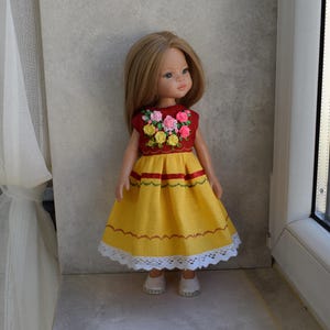Könnte beinhalten: Eine Puppe in einem gelben Kleid mit einem roten Oberteil, das mit bunten Blumen bestickt ist. Das Kleid hat rote und grüne Streifen und einen weißen Spitzensaum. Die Puppe hat blondes Haar und trägt weiße Schuhe.