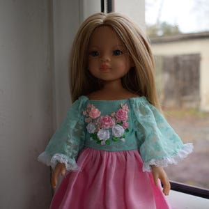 Könnte beinhalten: Eine Puppe mit langen blonden Haaren und blauen Augen, die ein rosa Kleid mit einem türkisfarbenen Oberteil trägt. Das Oberteil hat Spitzärmel und eine Blumenstickerei aus rosa und weißen Rosen.