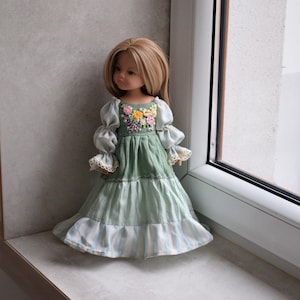 Könnte beinhalten: Puppe mit langem blondem Haar, bekleidet mit einem grünen Kleid mit floral besticktem Oberteil. Das Kleid hat lange Ärmel und einen gestreiften Rock. Die Puppe steht in der Nähe eines Fensters, mit einer neutralfarbenen Wand im Hintergrund.