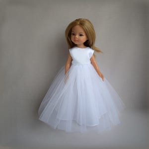 Könnte beinhalten: Eine Puppe mit langem hellbraunem Haar trägt ein ärmelloses weißes Kleid. Das Kleid besteht aus einem schlichten weißen Oberteil und einem voluminösen, mehrlagigen Tüllrock, der bis zum Boden reicht. Die Puppe hat einen neutralen Gesichtsausdruck und blickt nach vorne.