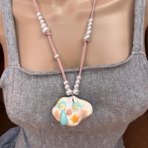 May include: A necklace featuring a pink cord strung with white faux pearls and a central ceramic pendant shaped like a cloud. The pendant has a glossy finish, a painted face with a blue teardrop, and a small yellow star accent on its surface.