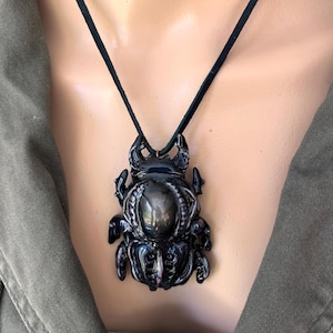 May include: A dark, iridescent beetle pendant necklace. The pendant is a detailed, sculpted insect with a large, oval-shaped center stone. The necklace has a black cord.