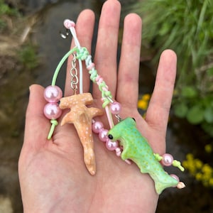 May include: Two handmade shark keychains. One is a hammerhead shark in peach with white spots, the other is a green shark with black eyes. Both have pink pearl beads and colorful cords.