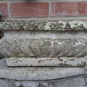 Op de afbeelding: Een rechthoekige stenen plantenbak met een verweerde, gebroken witte afwerking. De buitenkant is voorzien van een herhalend schulppatroon langs de basis en een gestructureerde, decoratieve rand aan de bovenkant, met tekenen van ouderdom.