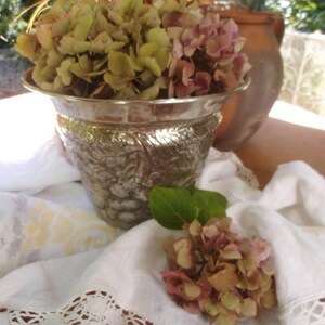 Può includere: Un vaso in metallo argentato, con rilievi, con un bouquet di ortensie verdi e rosa. Il vaso è appoggiato su una tovaglia bianca con pizzo.