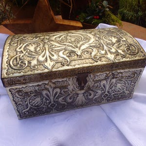 Puede incluir: Un cofre del tesoro de metal plateado con un diseño ornamentado. El cofre tiene una tapa con bisagras y una cerradura decorativa.