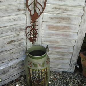 Op de afbeelding: Een olijfgroene metalen melkkan met een bosscène en een vogel erop geschilderd. De kan heeft een decoratieve rand en handvatten. Een roestige metalen blad- en spiraaldecoratie hangt boven de kan. De achtergrond is een witte, verweerde houten deur.