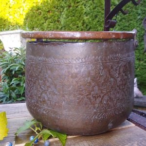 Op de afbeelding: Een grote koperen pot met een sierlijk, gegraveerd ontwerp. De pot heeft een koperen rand en een handvat. De pot staat op een houten oppervlak.