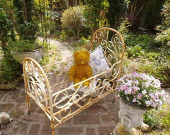 Könnte beinhalten: Ein weißes Metallbettchen mit einem gelben Teddybären, der darin sitzt. Das Bettchen ist mit kunstvollen Schnörkeln verziert und von grünem Laub umgeben.