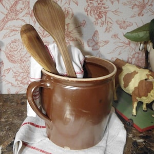 Può includere: Un vaso di ceramica marrone con un asciugamano a strisce bianche e rosse e due cucchiai di legno all'interno. Il vaso è appoggiato su un piano di lavoro in marmo.