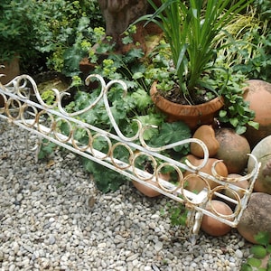 May include: A rectangular metal garden shelf featuring a white finish with visible rust accents. The structure includes a series of connected circular rings along the front edge and decorative scrollwork details along the top rail.