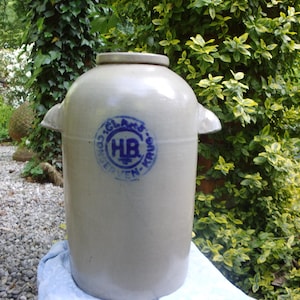 Può includere: Un grande boccale in gres bianco sporco con corpo arrotondato, due manici laterali e coperchio. Il boccale presenta un emblema circolare blu con testo. È appoggiato su un panno azzurro, con un lussureggiante sfondo verde.