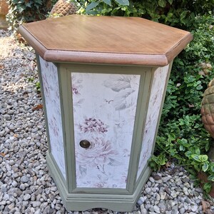 Peut inclure: Une table d'appoint hexagonale avec un plateau en bois naturel et un cadre peint en vert sauge. Les panneaux latéraux présentent un fond blanc avec un motif floral violet doux. Un petit bouton rond en métal foncé est centré sur la porte avant.