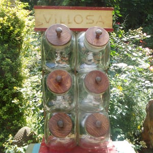 Könnte beinhalten: Vintage Glasbehälter-Display mit sechs durchsichtigen Glasbehältern, jeweils mit einem Holzdeckel und Knopf. Die Gläser sind in zwei Reihen zu je drei angeordnet und stehen auf einem roten Holzregal. Das Schild oben zeigt "VILLOSA" in verblichener Schrift.
