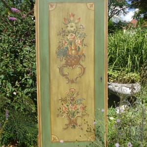 Op de afbeelding: Een vintage houten deur met een groen frame en een centraal paneel beschilderd met een bloemenontwerp. Het paneel toont een boeket bloemen en fruit in een mand, met een kleinere bloemstuk eronder. De deur staat tegen een achtergrond van weelderig groen.