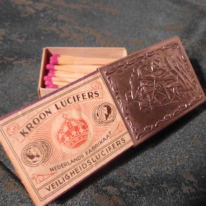 May include: A vintage matchbox with a red, white, and blue label that says "Kroon Lucifers" and "Nederlands Fabrikaat Veiligheidslucifers". The matchbox has a silver metal cover with an embossed design.