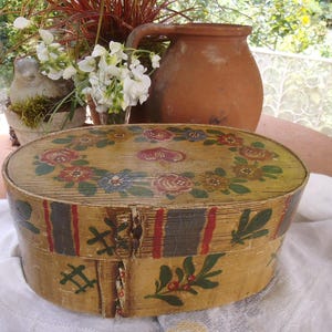 Può includere: Una scatola di legno vintage con un design floreale dipinto a mano. La scatola è di forma ovale e ha un coperchio. I fiori sono dipinti in tonalità di rosso, giallo, blu e verde. La scatola è appoggiata su un panno bianco.