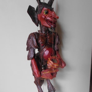 May include: A red and black wooden puppet of a devil with horns, a long tail, and a mischievous expression. The puppet is suspended by strings and has a red and gold painted hand.