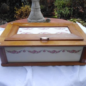 Può includere: Scatola portagioie in legno con finitura marrone chiaro. La scatola presenta un motivo floreale decorativo in rosa e bianco sulla parte anteriore e superiore. Il coperchio ha un pannello di vetro con un design floreale abbinato. Un piccolo fermaglio in metallo fissa il coperchio.