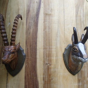 以下が含まれることがあります： 木製の動物の頭部の壁掛けが2つ。左側の壁掛けは、茶色と黒のヤギの頭で、長く曲がった角がついている。右側の壁掛けは、茶色と白の鹿の頭で、短く曲がった角がついている。
