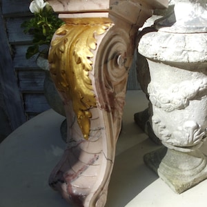 May include: A marble corbel with a gold leaf design. The corbel has a rectangular top and a curved, ornate base. The color is a mix of pink, white, and gold. The corbel is next to a white urn.