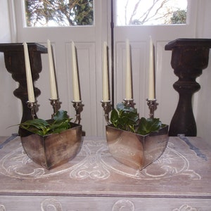 Könnte beinhalten: Zwei silberne Kerzenhalter mit weißen Kerzen und grünen Pflanzen in silbernen Töpfen auf einer hellbraunen Holzoberfläche mit geschnitzten Verzierungen.