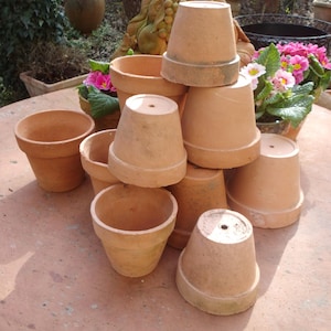 Könnte beinhalten: Eine Ansammlung von Terrakotta-Blumentöpfen in verschiedenen Größen, einige gestapelt. Die Töpfe haben ein klassisches, konisches Design und einen warmen, erdigen Farbton. Rosa Blüten sind im Hintergrund zu sehen.